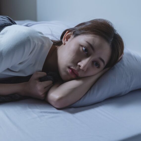 A person lies on their side in bed, resting their head on a pillow and looking off into the distance with a thoughtful expression.