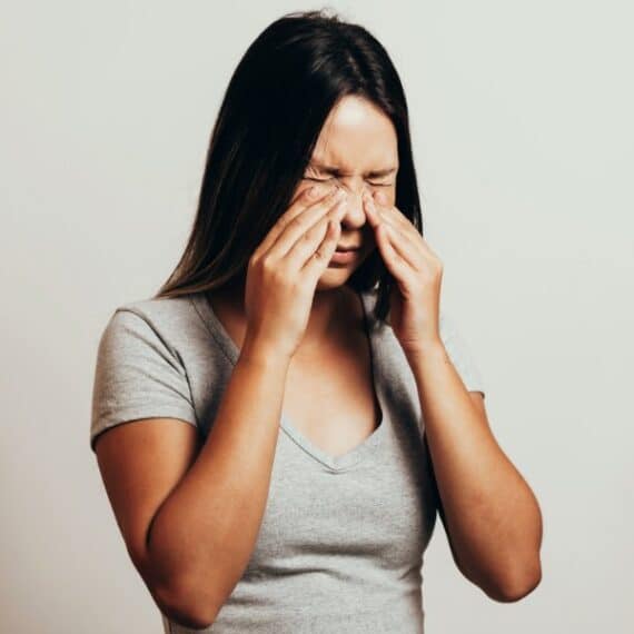 A woman with long dark hair wearing a gray shirt rubs her eyes with both hands, standing against a plain light background.