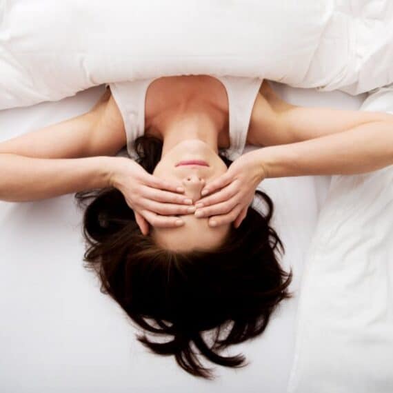 A person lies on a bed under a white blanket, covering their eyes with both hands.
