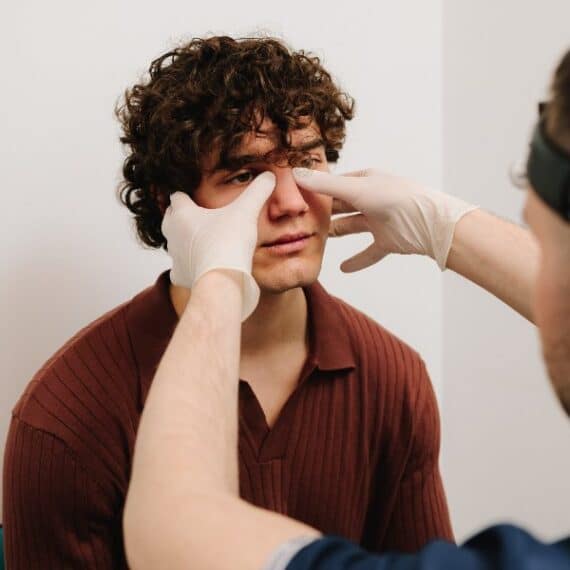 A healthcare professional wearing gloves examines a seated man's eyes by gently touching the area below his eyes in a clinical setting.