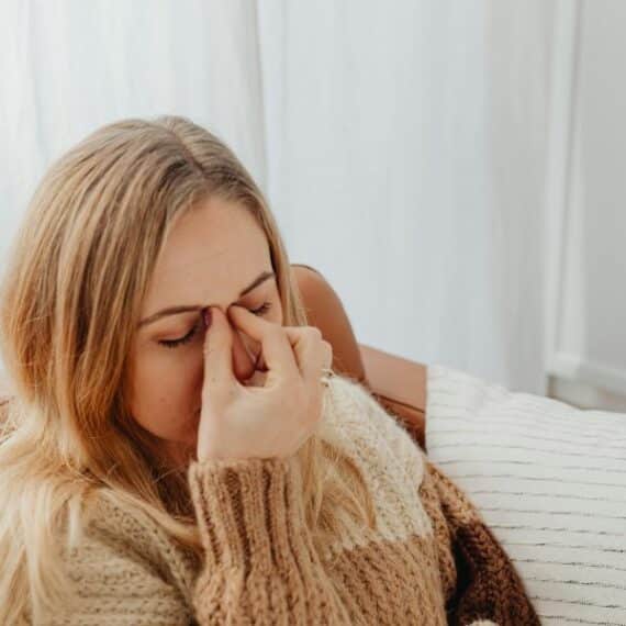 A woman in a sweater sits on a couch with her eyes closed, pinching the bridge of her nose, appearing tired or stressed—an expression often seen when to suspect chronic sinusitis.